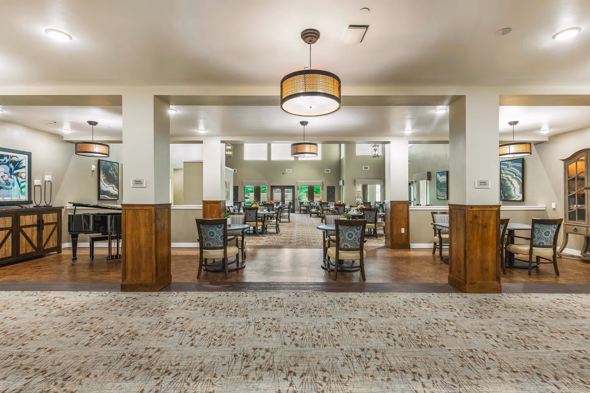 Spacious and well-lit dining area in a senior living facility with multiple round tables and chairs arranged neatly. The room features wooden paneling on the lower walls, modern hanging light fixtures, framed artwork, and a piano on the left side. Large windows at the far end allow natural light to brighten the space.