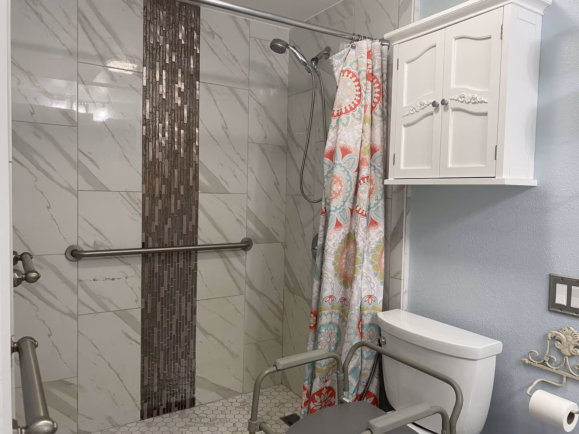 A bathroom featuring a walk-in shower with a vertical strip of decorative tiles, a colorful floral shower curtain, grab bars for safety, a white wall-mounted cabinet above the toilet, and a toilet with an attached shower chair.