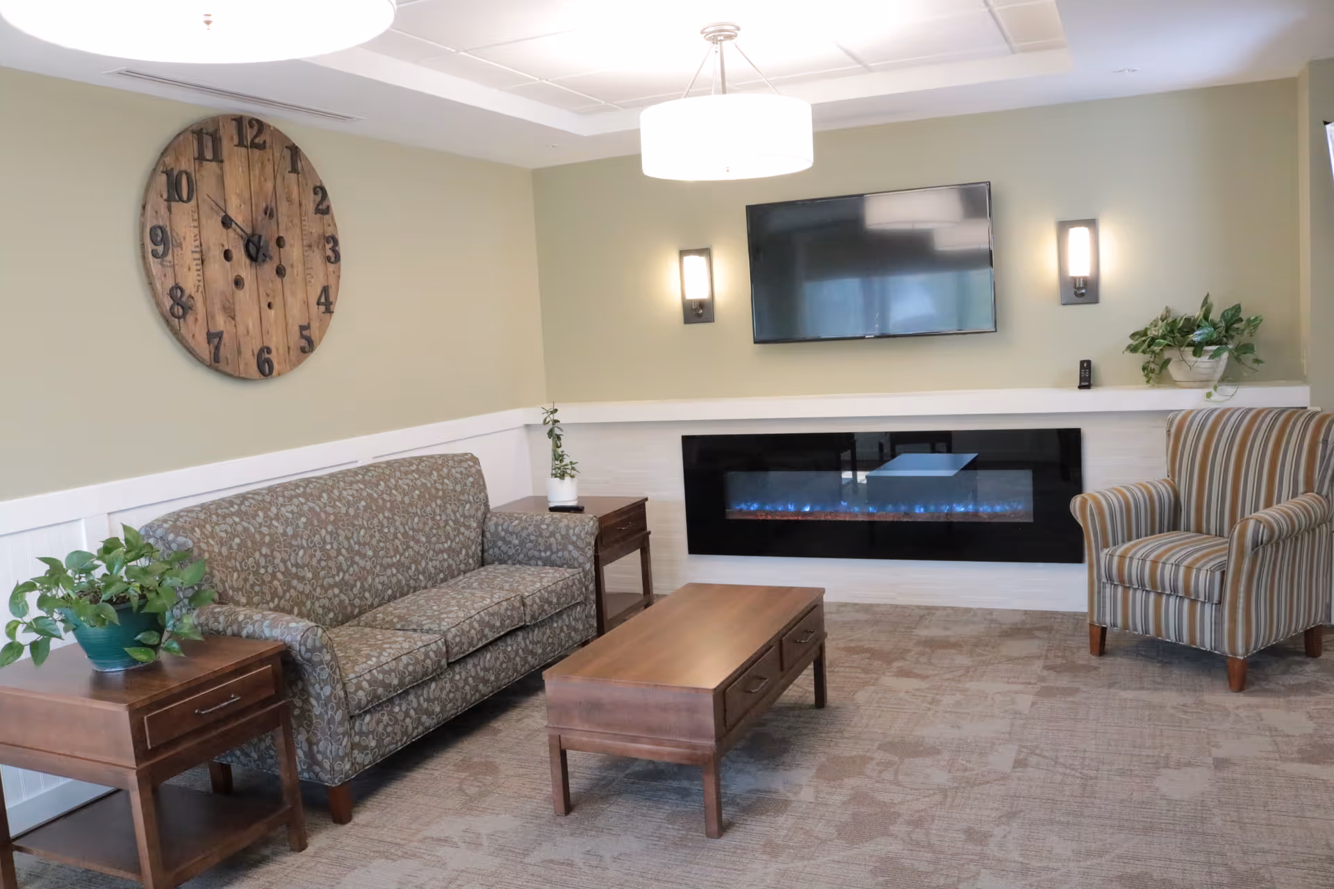 A cozy living room with a patterned sofa, a striped armchair, a wooden coffee table, and two side tables with potted plants. A large wooden clock hangs on the wall above the sofa. A flat-screen TV is mounted above a modern electric fireplace, flanked by two wall sconces. The room has soft green walls and carpeted flooring.