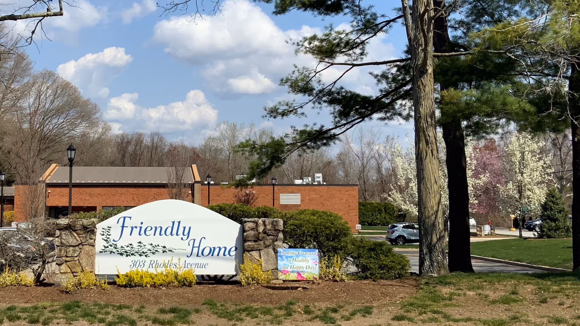 Outdoor view of the entrance sign for Friendly Home at 303 Rhodes Avenue, surrounded by stone pillars, bushes, trees, and a clear blue sky with some clouds.