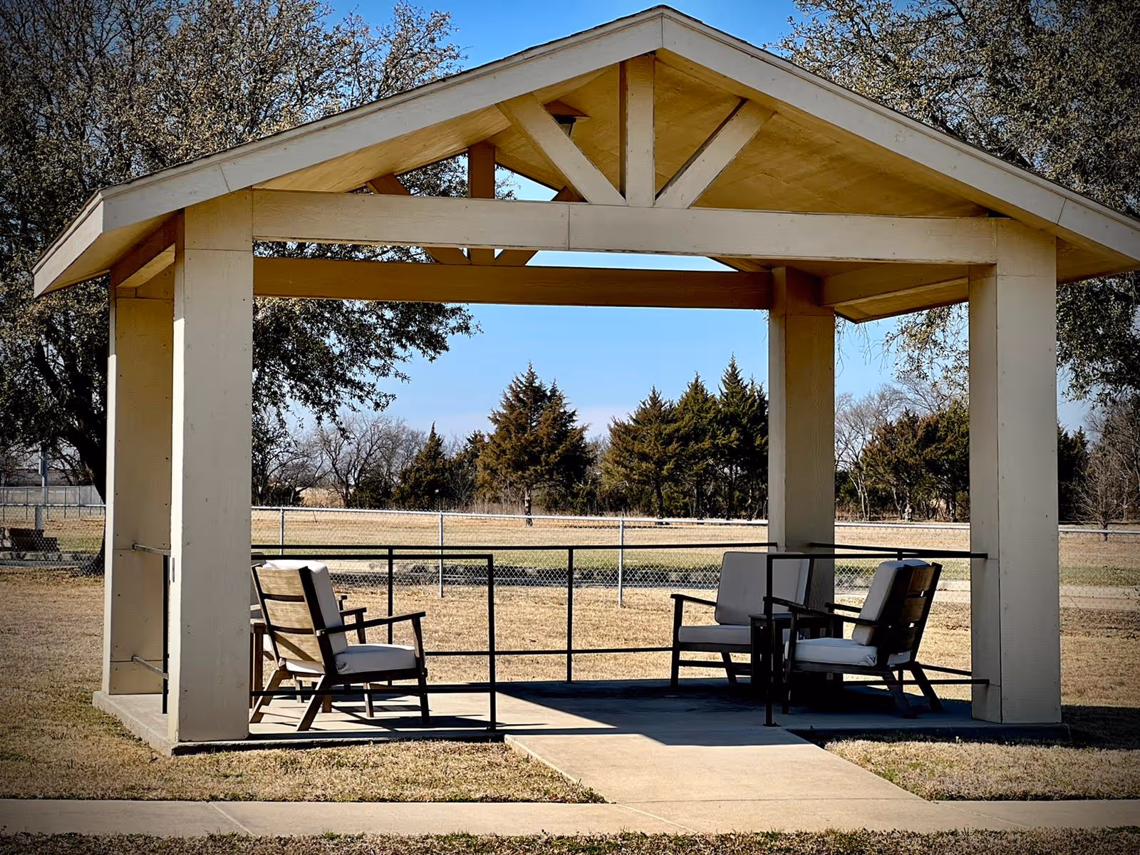 A wooden gazebo with a peaked roof situated outdoors on a grassy area. Inside the gazebo, there are four cushioned chairs arranged around a small table. Trees and a chain-link fence are visible in the background under a clear blue sky.