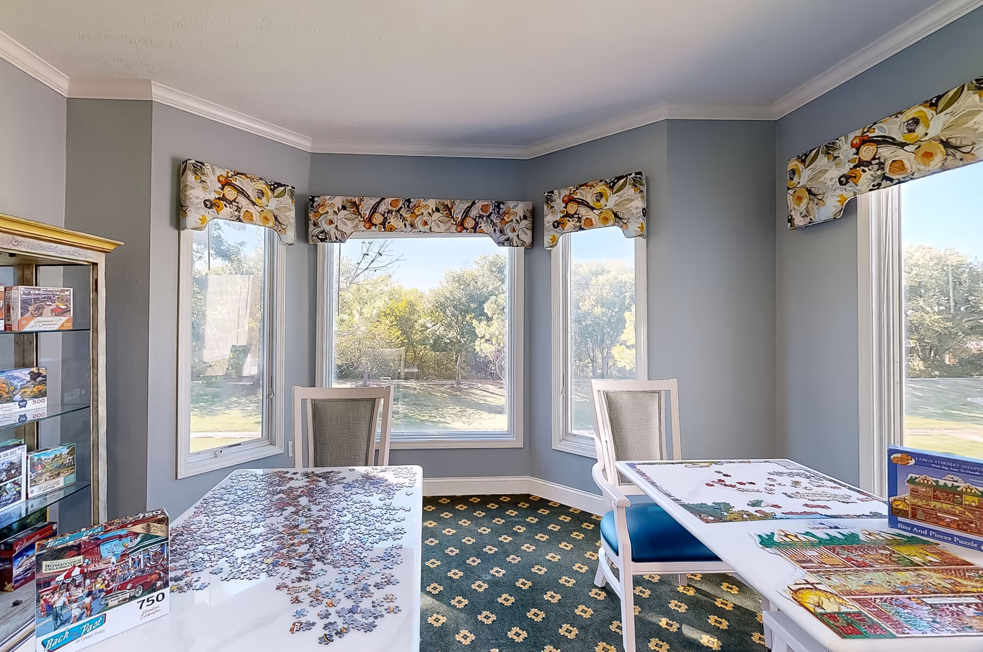 A bright room with large windows showing a green outdoor view. Two tables with partially completed jigsaw puzzles are placed in the room, each accompanied by a chair. The walls are painted blue-gray, and the windows have floral valances. A glass cabinet with more puzzles is visible on the left side.