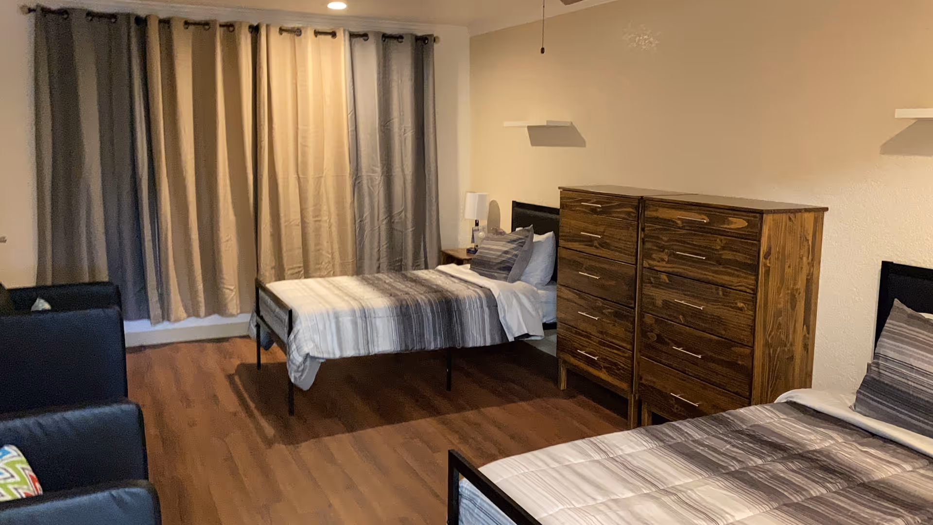 A bedroom with two single beds, each with striped bedding and pillows. There are two wooden dressers with multiple drawers between the beds. The room has wood flooring, beige walls, and a window covered with beige and gray curtains. Two black armchairs with colorful pillows are positioned on the left side of the room.