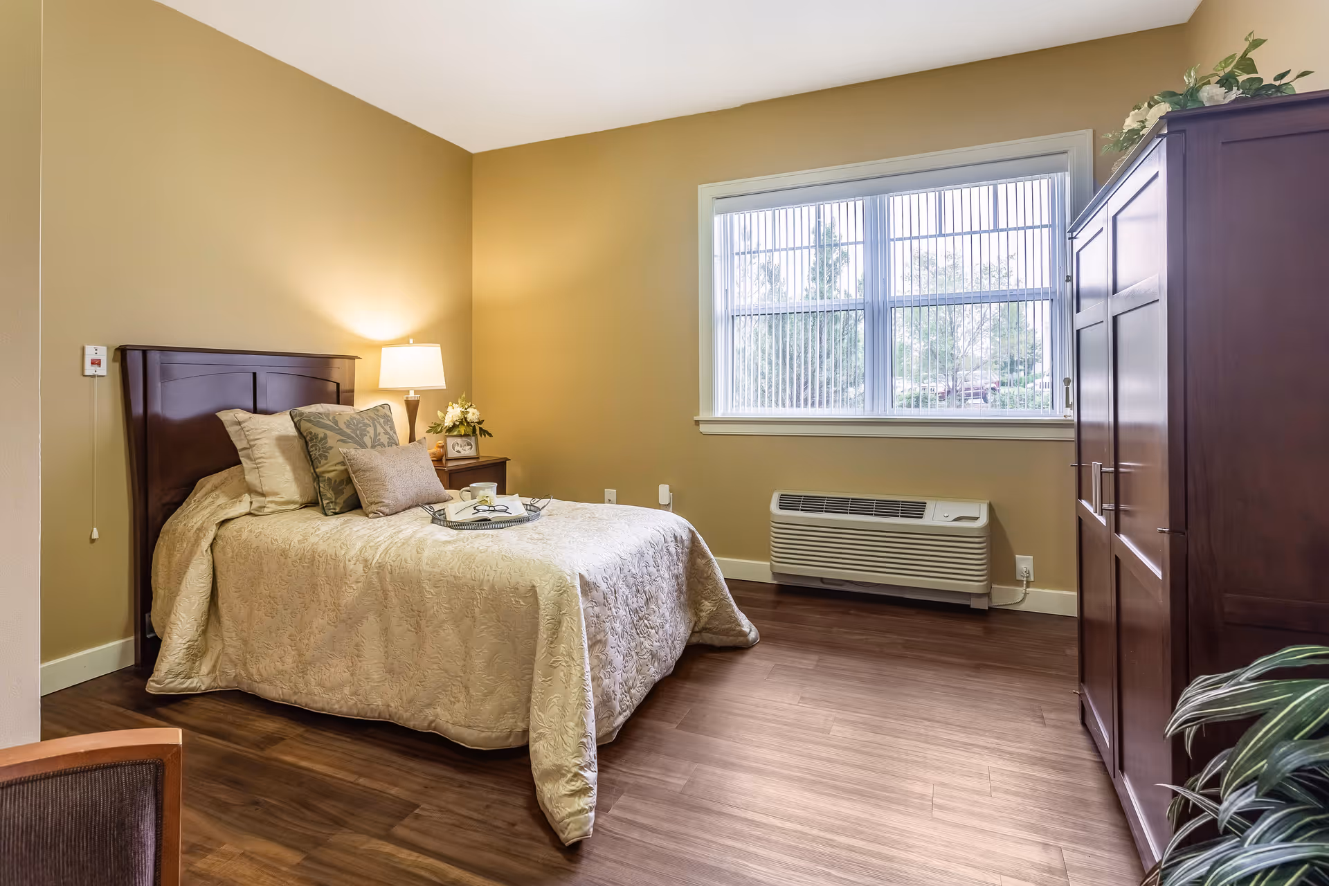 A cozy bedroom with a neatly made bed featuring beige bedding and multiple pillows. There is a wooden headboard, a nightstand with a lamp and flowers, a large window with vertical blinds, a wooden wardrobe, and a heating/cooling unit below the window. The room has warm beige walls and wooden flooring.
