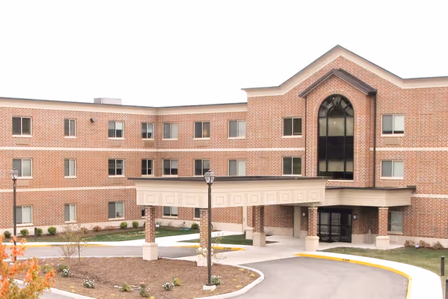 Exterior view of a three-story brick senior living facility with multiple windows, a covered entrance with columns, a curved driveway, and landscaped areas with small plants and trees.