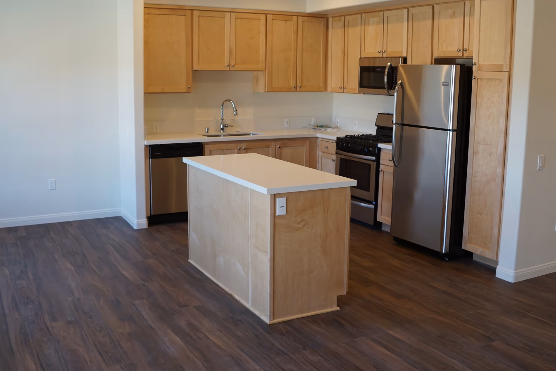 Modern kitchen with light wood cabinets, stainless steel refrigerator, stove, microwave, dishwasher, and a central island with a white countertop. The floor is dark wood and the walls are light-colored.