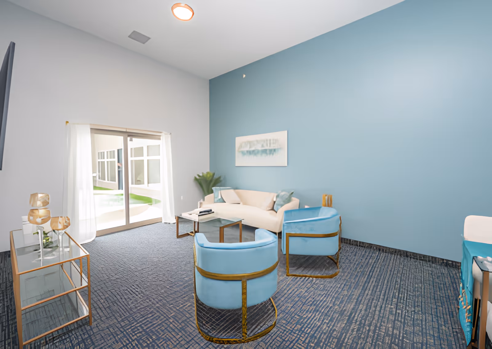 A modern living room with a light blue accent wall, a beige sofa with cushions, two light blue armchairs with gold frames, a glass coffee table, and a glass side table with decorative items. There is a large sliding glass door with white curtains letting in natural light and a view of an outdoor area.