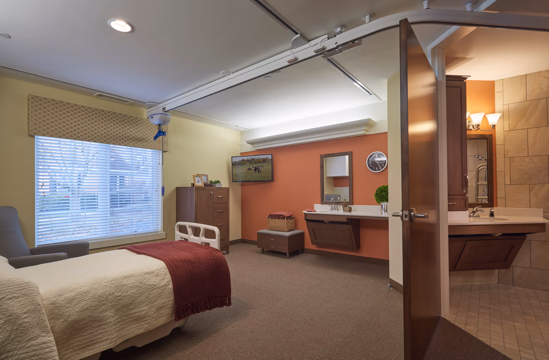 A well-lit bedroom in a senior living facility with a hospital-style bed covered with a cream blanket and a red throw. There is a large window with blinds and a patterned valance, a comfortable armchair, a wooden dresser, a wall-mounted TV, and a vanity area with a mirror and sink. An open door reveals a bathroom with a sink and tiled walls.