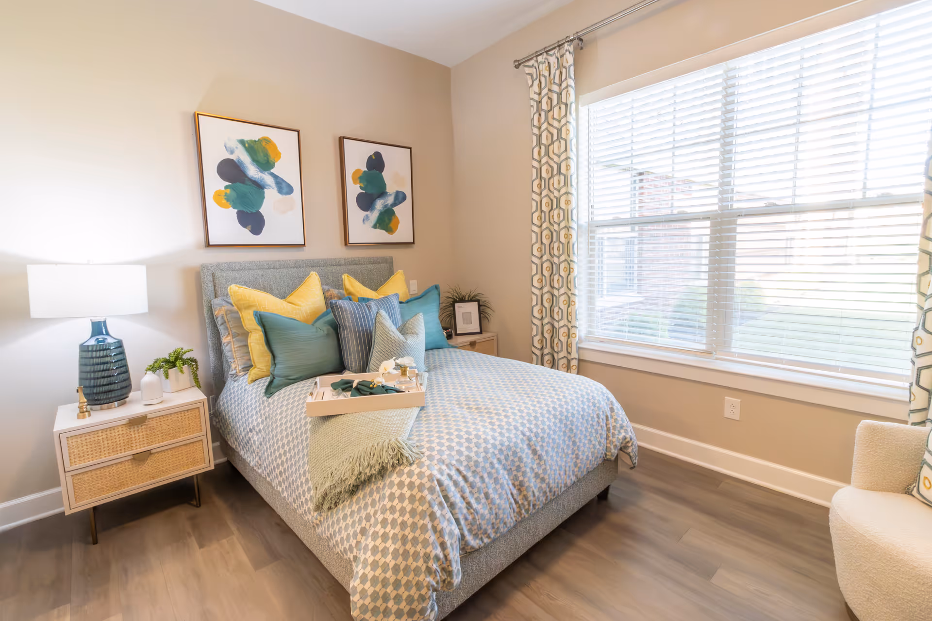 Sunlit furnished bedroom with a made bed, decorative pillows, a nightstand with a lamp, wall art, and a large window with blinds.