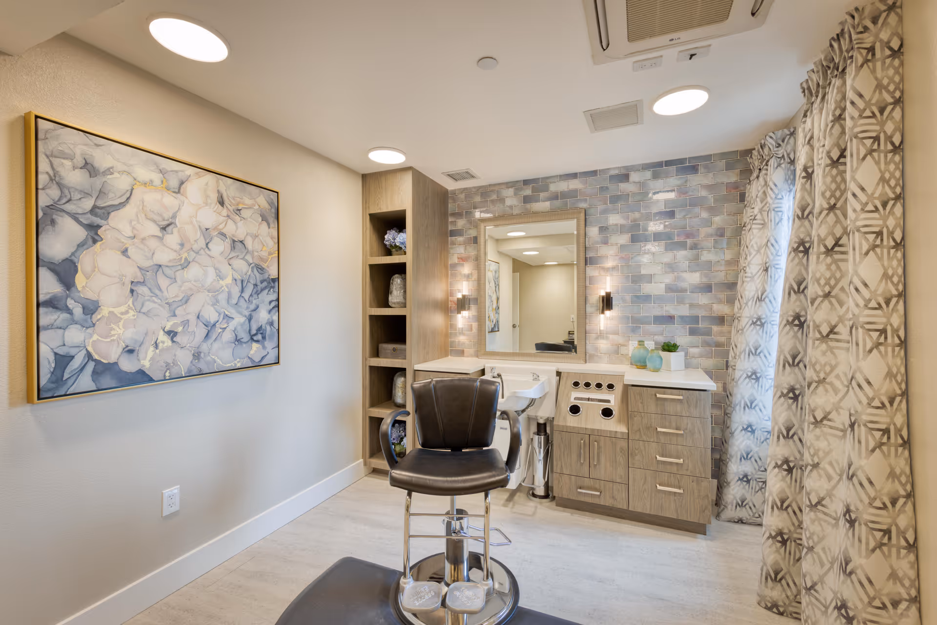 Well-lit hair styling station with a black salon chair in front of a mirror, sink and cabinetry against a tiled tile backsplash.