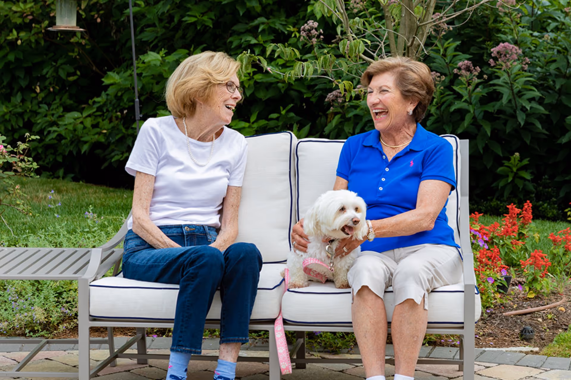 Two elderly women sitting on an outdoor cushioned bench in a garden, smiling and laughing together. One woman is wearing a white shirt and jeans, and the other is wearing a blue polo shirt and light-colored pants, holding a small white dog on her lap. There are green bushes and colorful flowers in the background.