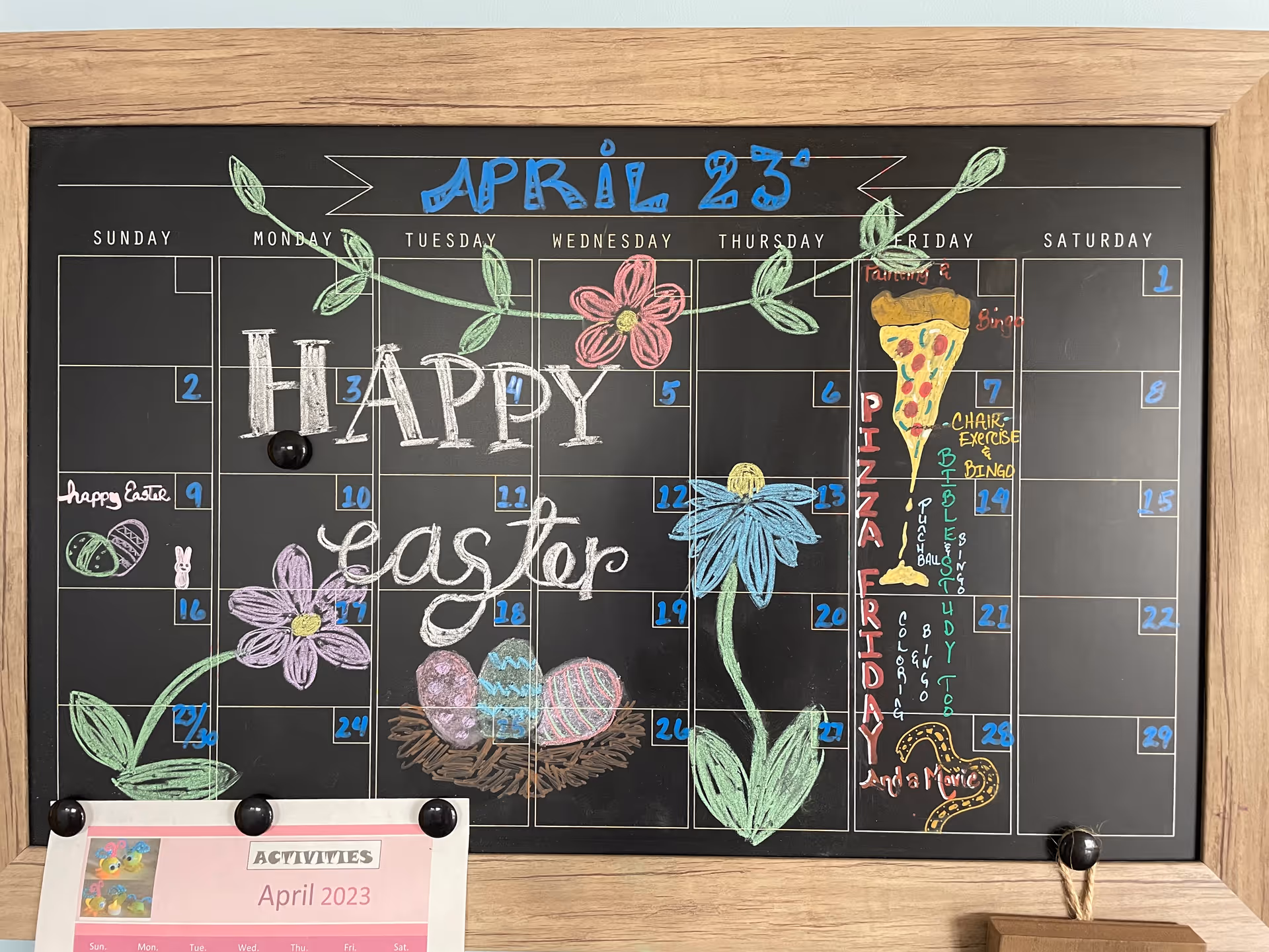 Chalkboard calendar for April 2023 in a wooden frame decorated with "Happy Easter" lettering, colorful flowers, painted eggs, and activity notes.