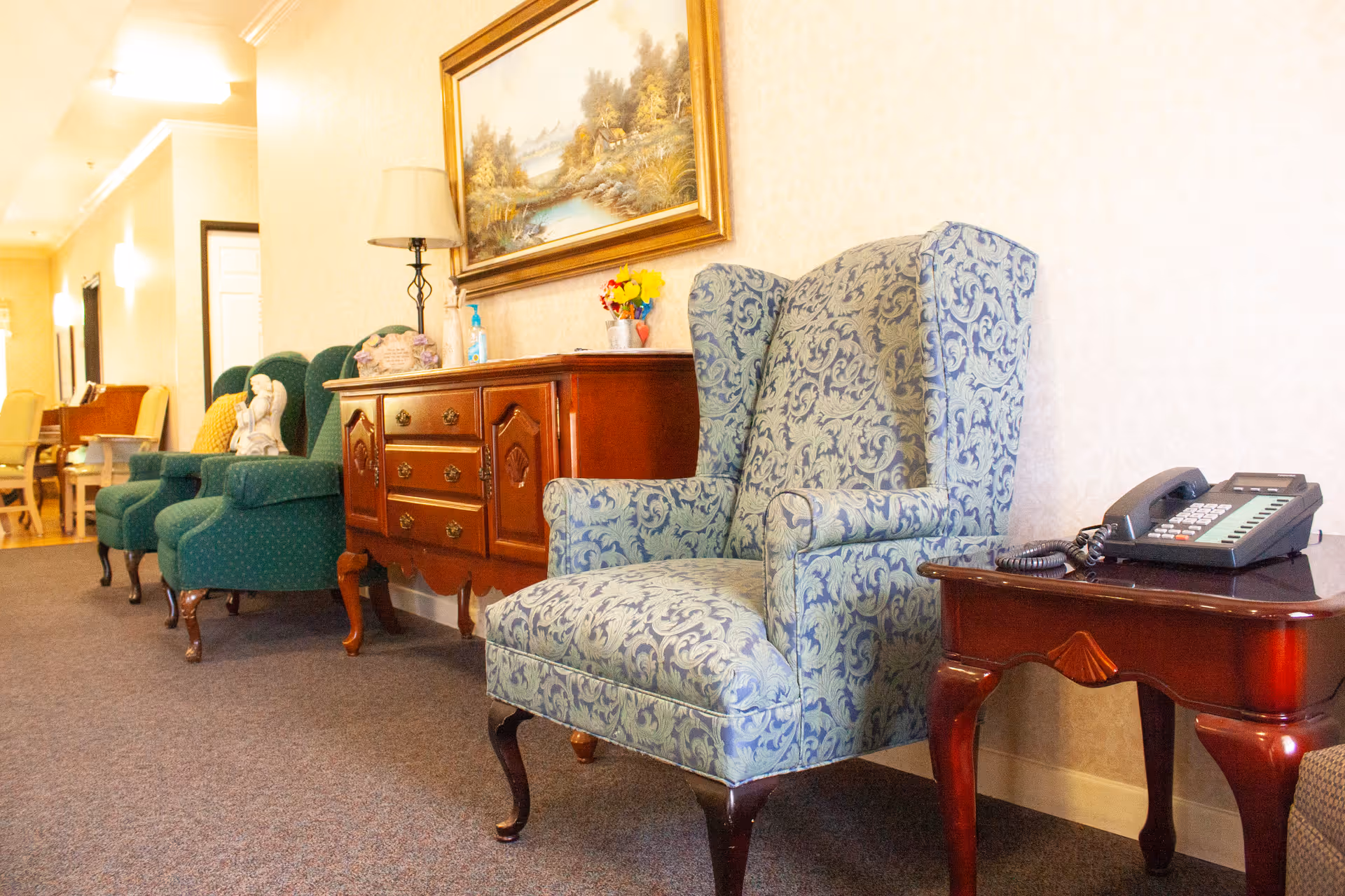 A row of upholstered wingback and armchairs with wooden side tables, a phone, lamp, and framed painting in a carpeted assisted-living hallway.