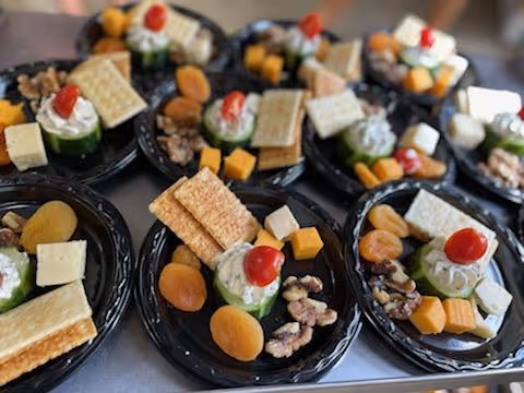 Several black appetizer plates arranged with crackers, cheese cubes, cucumber cups topped with cream and a cherry tomato, dried apricots, and walnuts.