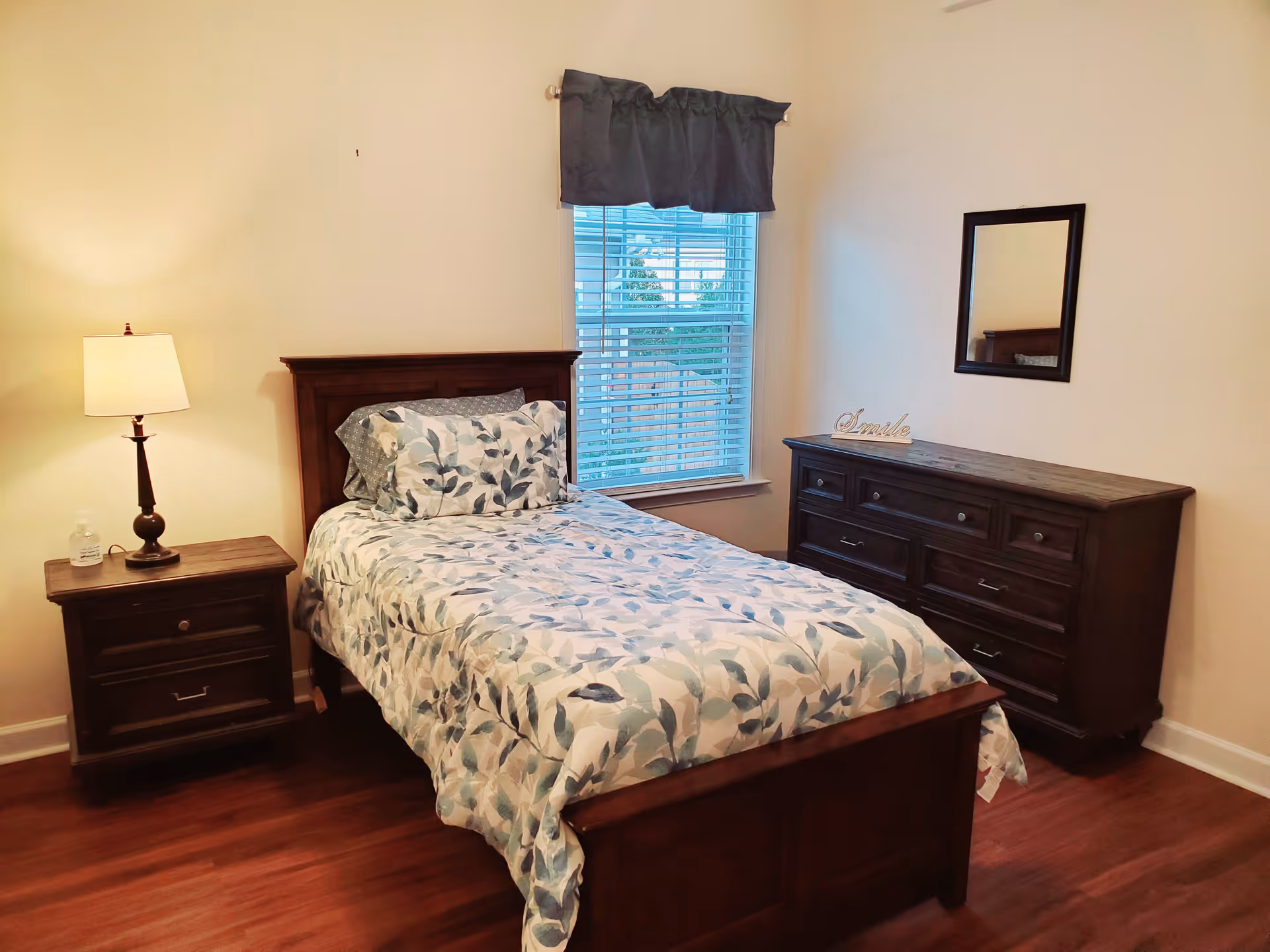 A neatly made single bed with a floral patterned comforter and pillows in a bedroom. Next to the bed is a wooden nightstand with a lamp and a bottle of hand sanitizer. Across from the bed is a wooden dresser with a mirror above it and a decorative 'Smile' sign on top. A window with blinds and a dark valance is centered on the wall behind the bed.