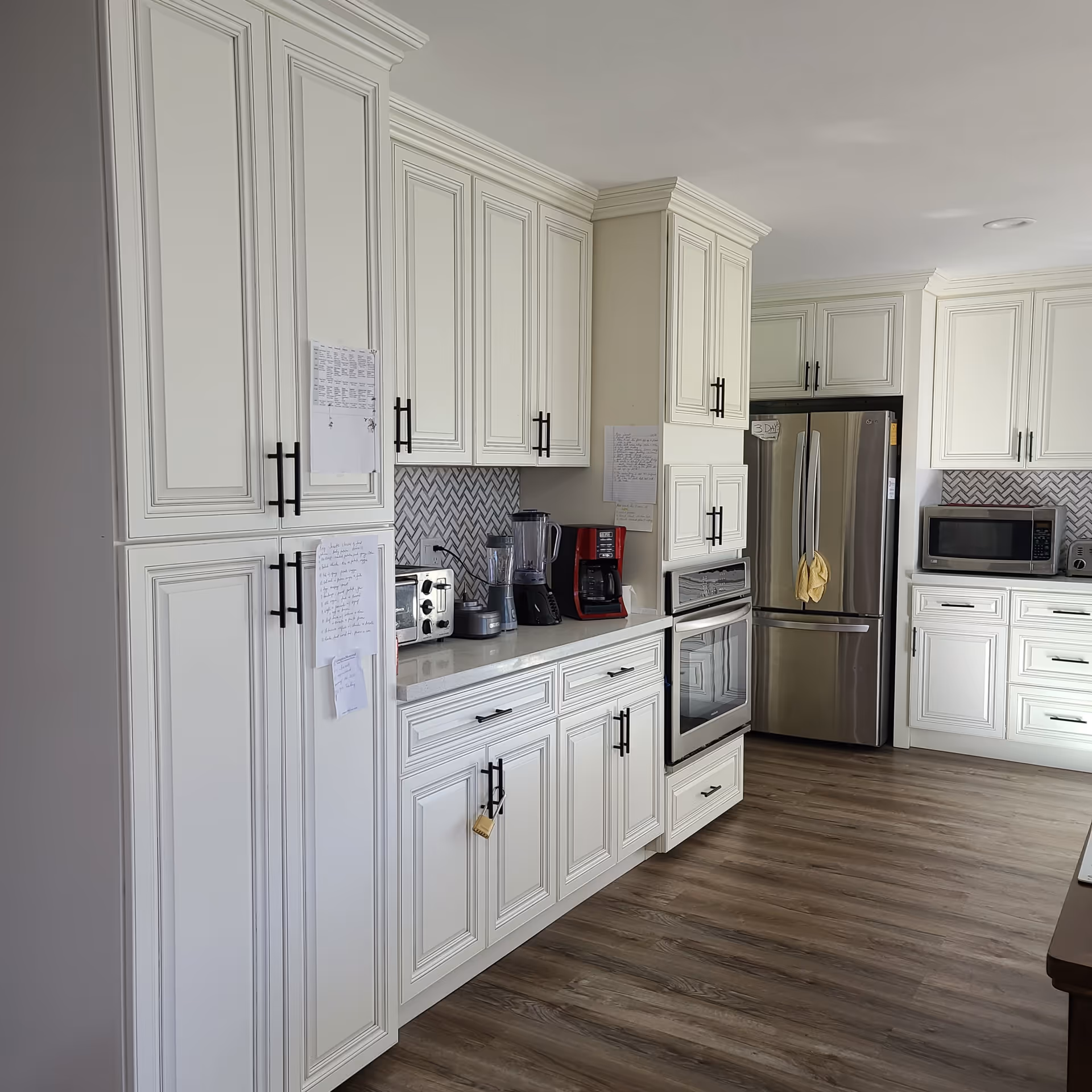 A modern kitchen with white cabinetry and black handles, featuring a stainless steel refrigerator, built-in oven, microwave, toaster, blender, and coffee maker. The backsplash has a herringbone tile pattern, and the floor is wood with a light finish. Several papers are attached to the cabinets.