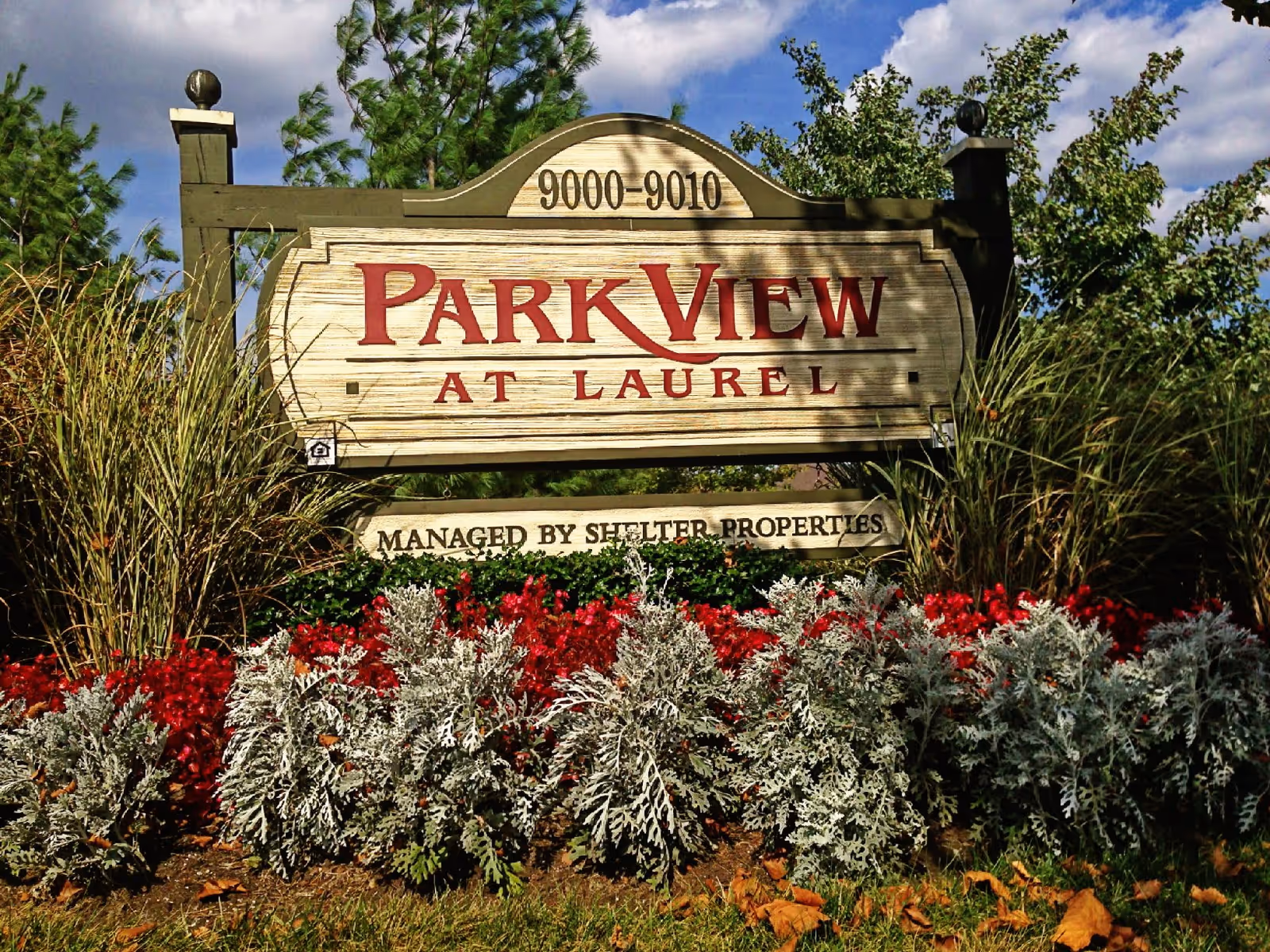 Outdoor wooden sign for Park View at Laurel I, surrounded by decorative plants and flowers, with a partly cloudy blue sky in the background.