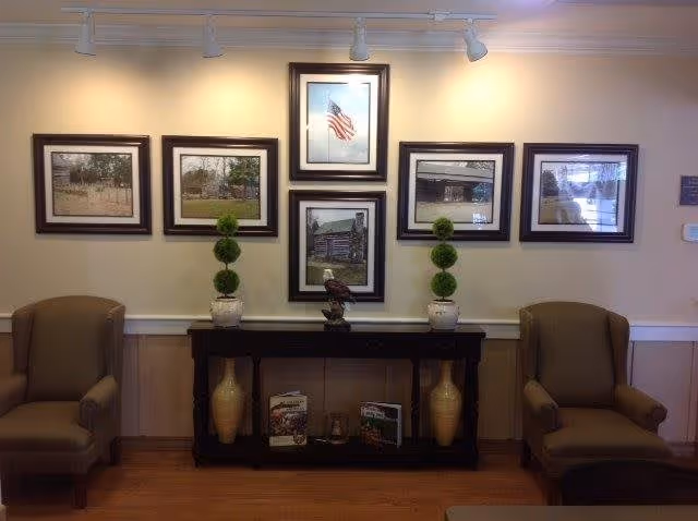 A cozy seating area with two brown armchairs on either side of a black console table. The table holds two potted topiary plants, a decorative eagle statue, two large vases, and some books. Above the table, six framed pictures are arranged on a beige wall, including an American flag and various outdoor scenes. The floor is wooden, and the lighting is provided by ceiling spotlights.