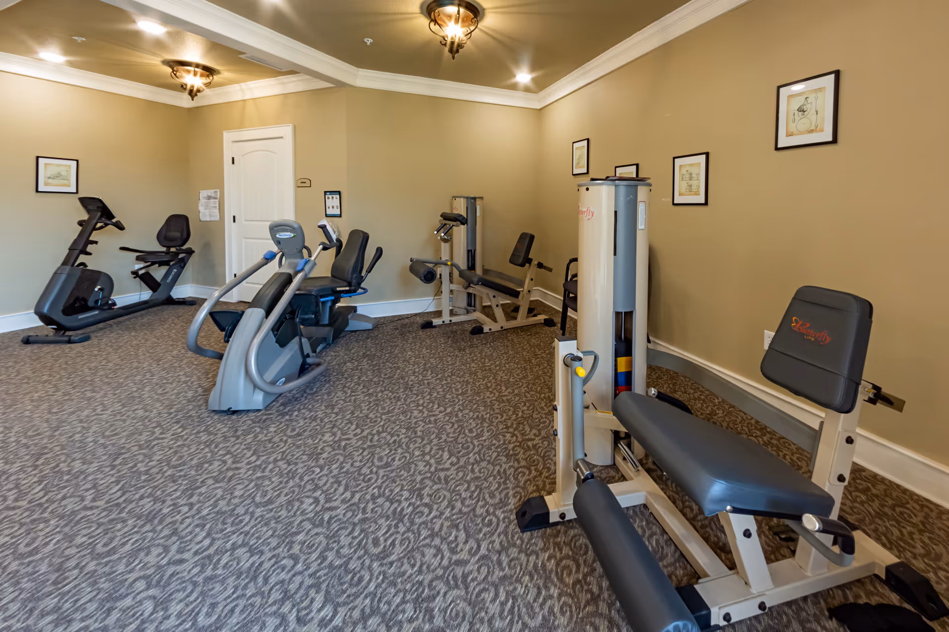 A fitness room with various exercise equipment including stationary bikes, a leg press machine, and other workout machines. The room has beige walls, patterned carpet, framed pictures on the walls, and ceiling lights.