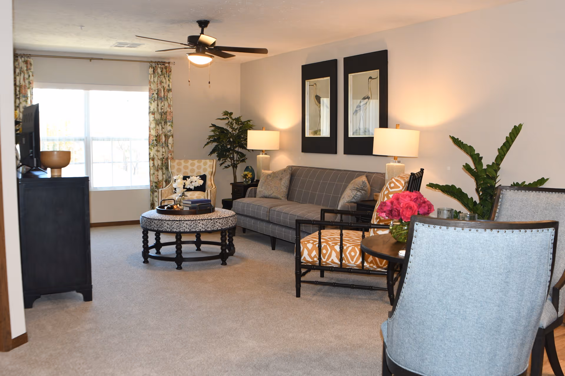 Furnished living room with a sofa, patterned chairs, round coffee table, TV console, lamps, and plants by a large window.