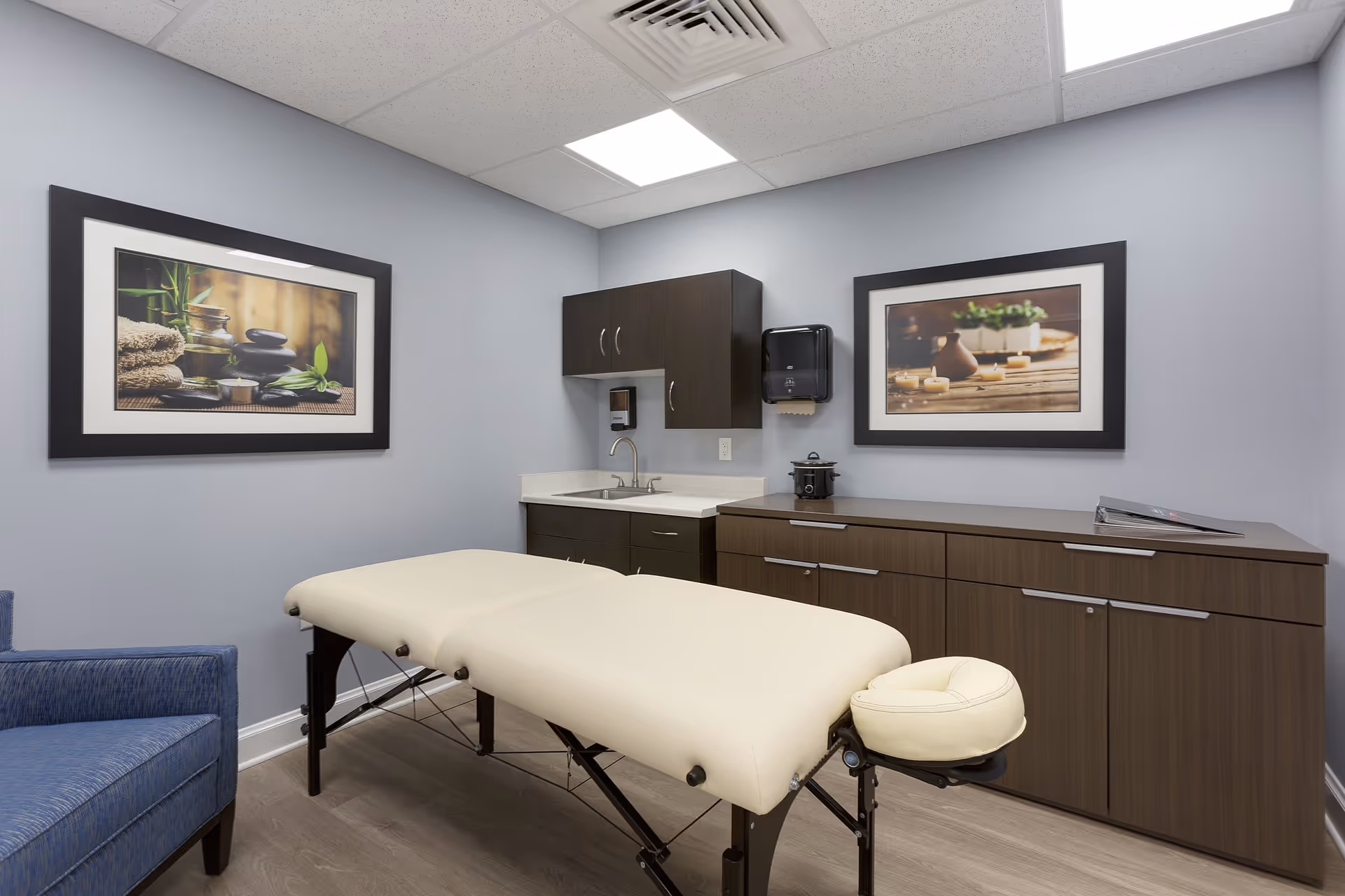 Massage treatment room with a cream-colored massage table, cabinetry, sink, and framed artwork on the walls.