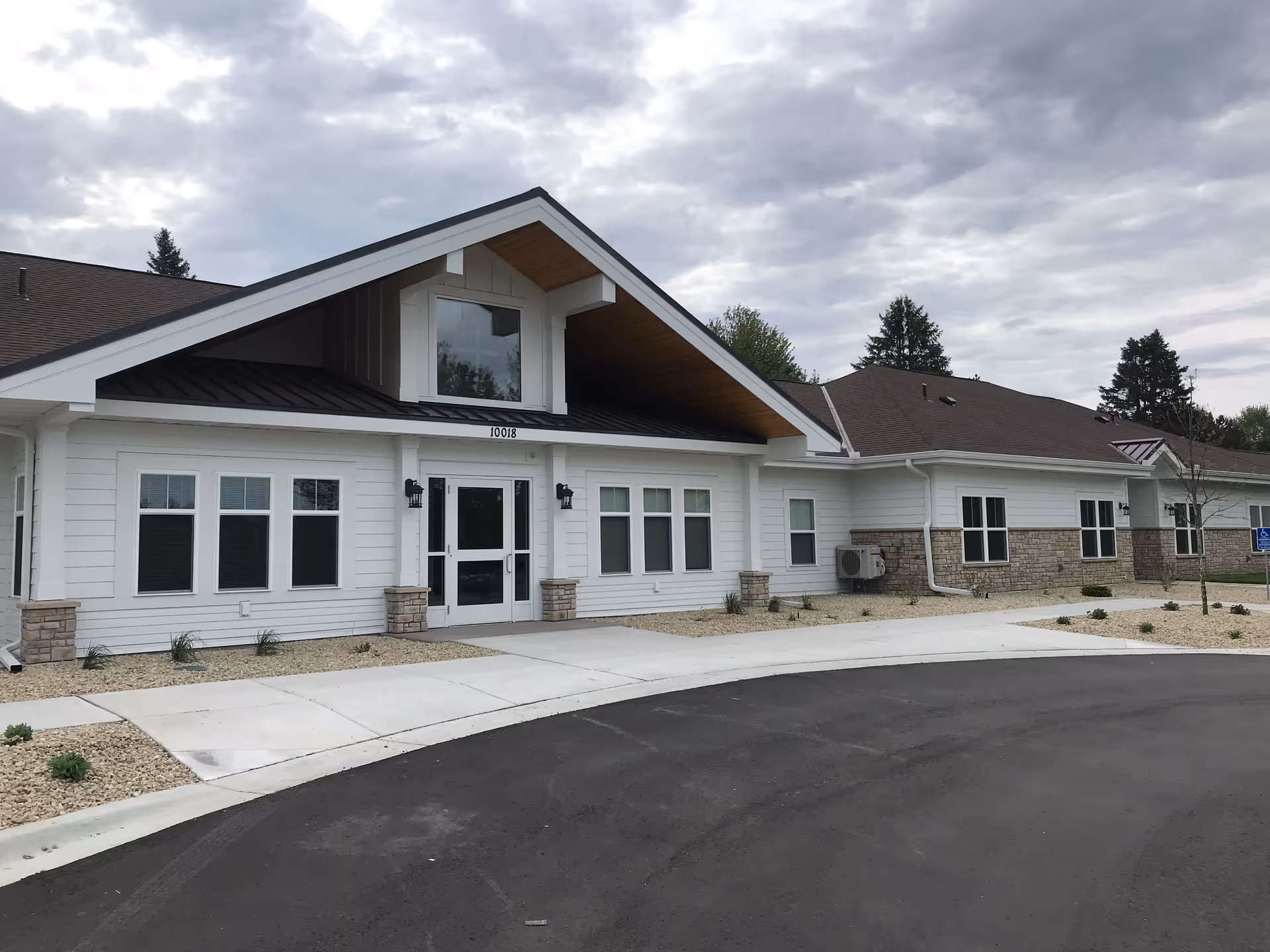 Front exterior of a single-story white building with a large gabled entrance and paved circular driveway under a cloudy sky.