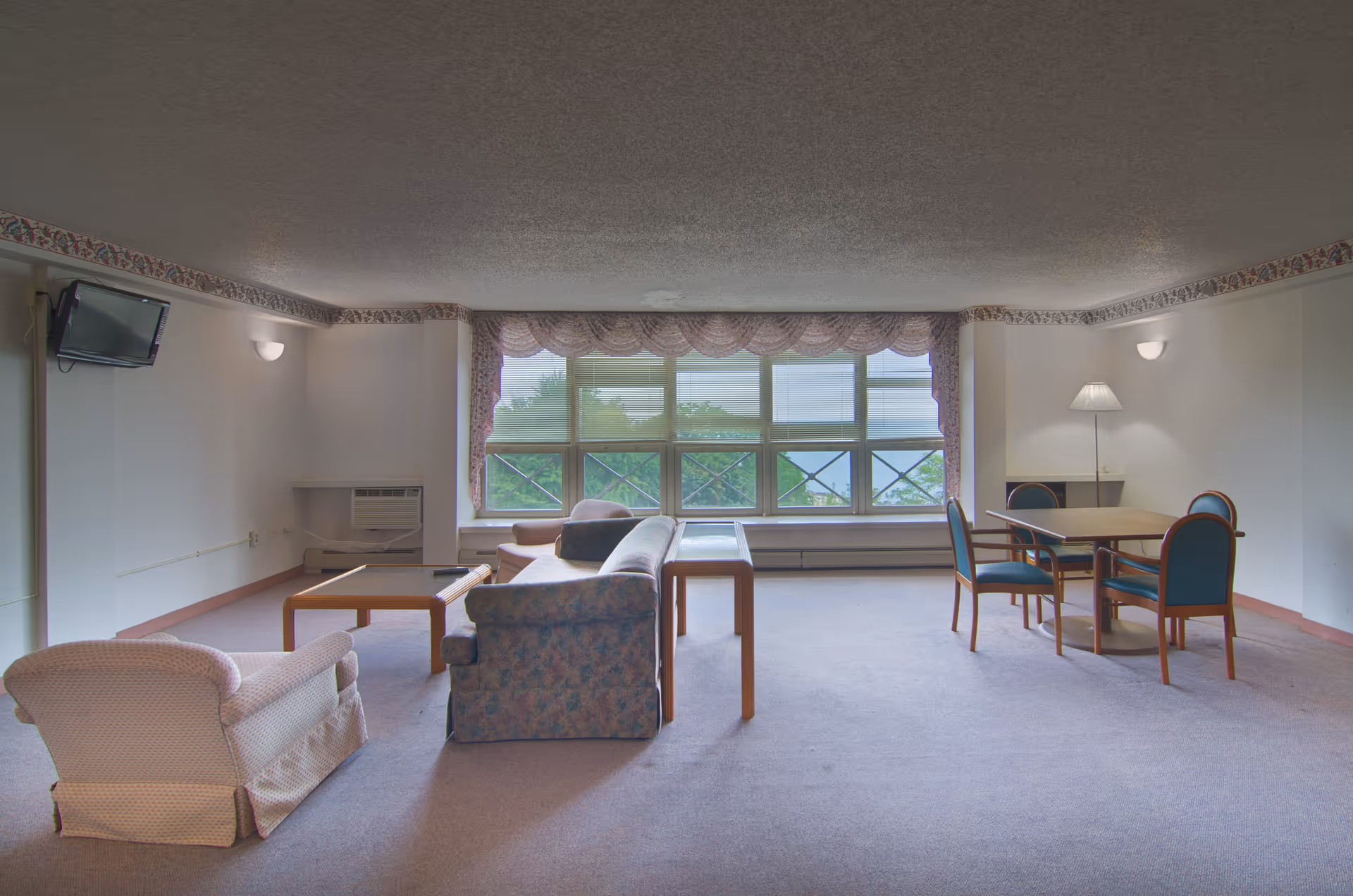 A spacious living room area with a large window covered by sheer curtains and valances, allowing natural light to brighten the room. The room contains a floral-patterned sofa, a cushioned armchair, a wooden coffee table, and a small dining table with four chairs. A wall-mounted TV is visible on the left side, and a floor lamp stands near the dining table. The walls are light-colored with a decorative border near the ceiling.