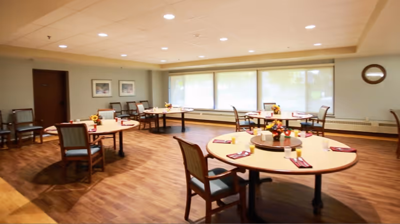 Dining room with round tables set with place settings and chairs on a wood floor near large windows.