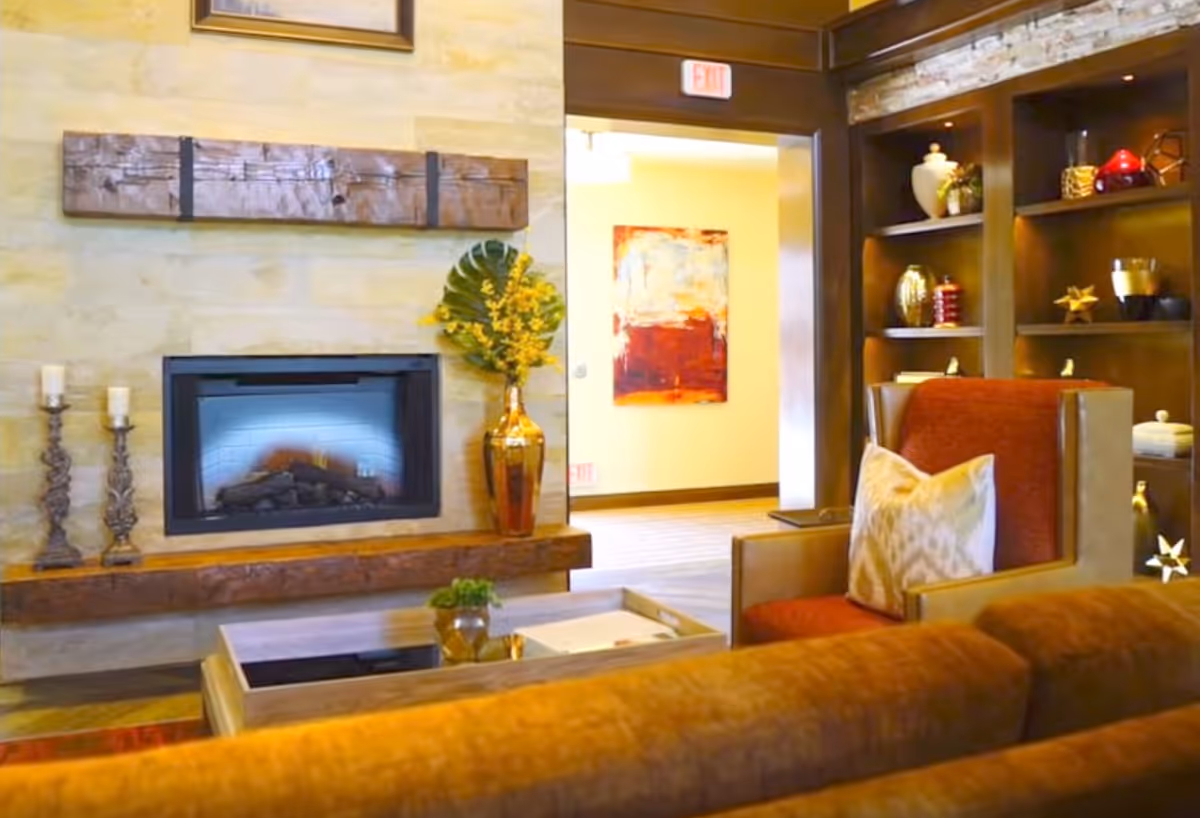 A cozy living room area featuring a modern fireplace set in a stone wall with two decorative candles on the left and a tall vase with yellow flowers on the right. In front of the fireplace is a wooden coffee table with a small plant and a tray. To the right, there is a comfortable armchair with a patterned pillow, and behind it, built-in wooden shelves display various decorative items. An open doorway reveals a brightly lit hallway with a colorful abstract painting on the wall.