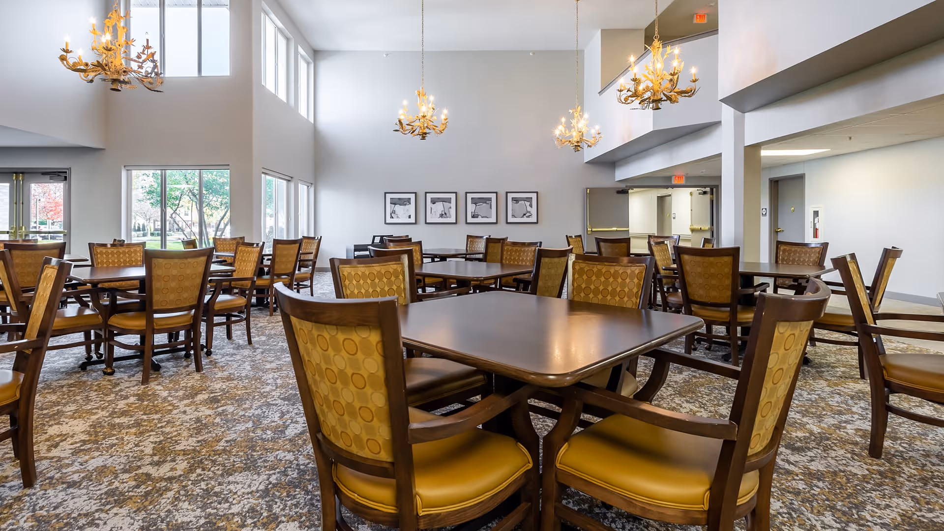 A spacious dining room with multiple wooden tables and chairs featuring yellow patterned upholstery. The room has large windows allowing natural light to fill the space, and several ornate chandeliers hanging from the ceiling. The carpet has a patterned design, and there are framed black and white pictures on the far wall.