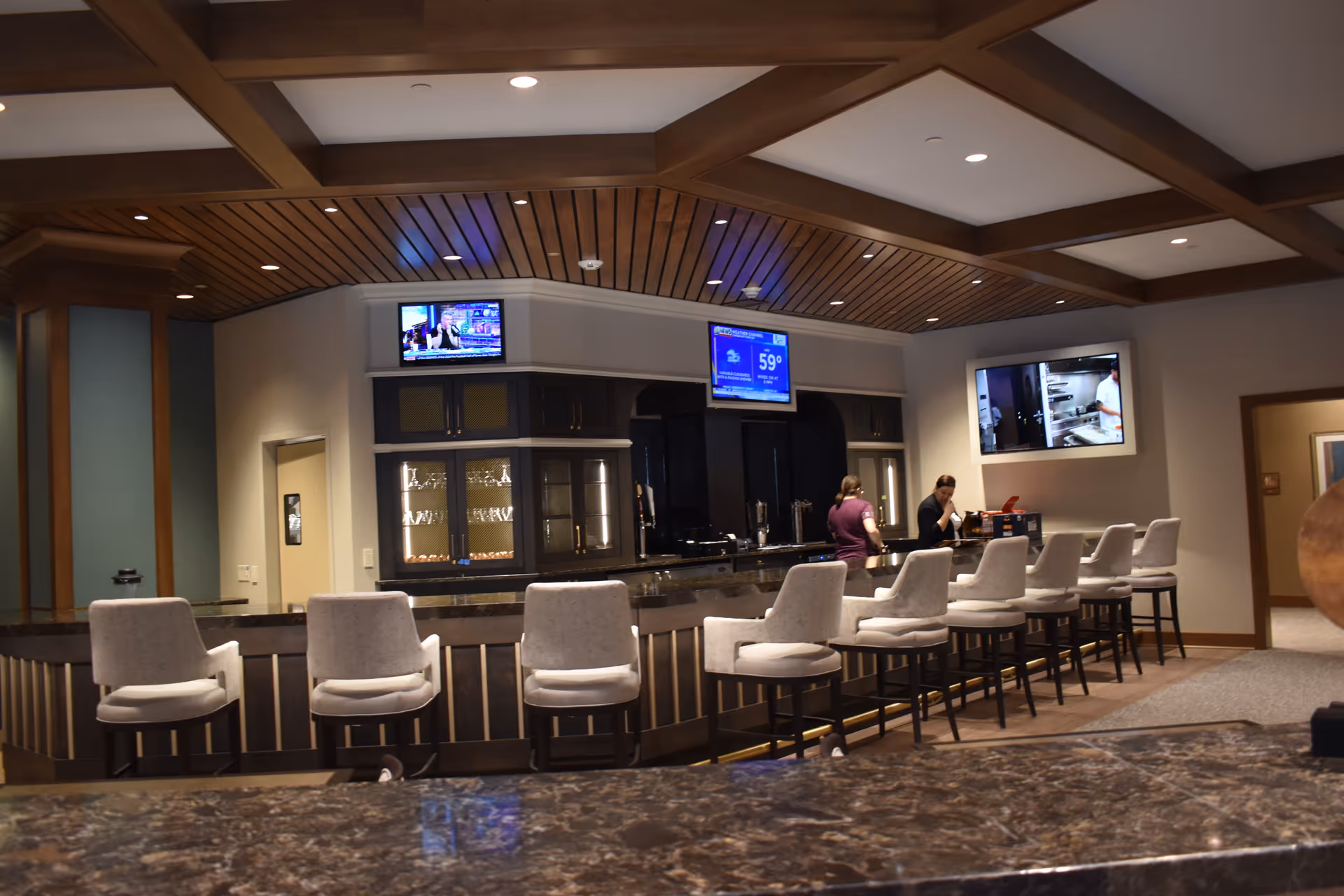Interior view of a modern bar area with a row of cushioned bar stools along a counter. Two people are behind the counter, and three televisions are mounted on the walls above the bar. The ceiling has wooden beams and recessed lighting.