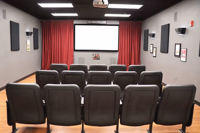 A small theater room with three rows of black cushioned seats facing a large white projection screen framed by red curtains. The walls are gray with black acoustic panels and framed pictures, and the ceiling has recessed lighting and a projector mounted.