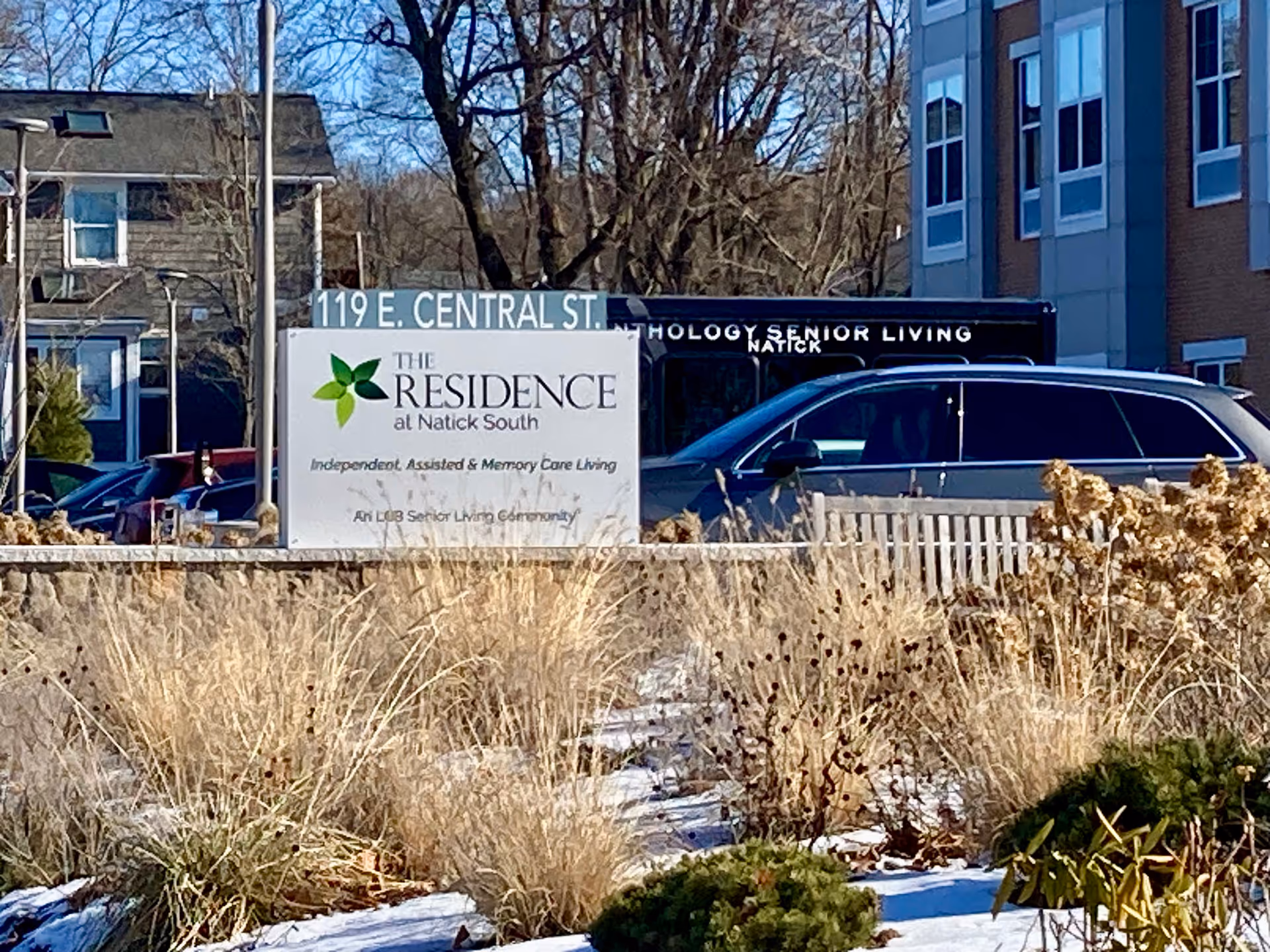 Outdoor view of The Residence at Natick South sign in front of the facility with winter landscaping and parked cars.