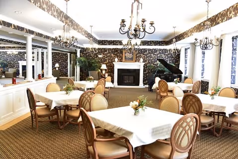 A bright and elegant dining room with several tables covered in white tablecloths, each adorned with small floral centerpieces. The room features patterned wallpaper, multiple chandeliers, a fireplace, a grand piano, and large windows with curtains allowing natural light to fill the space.