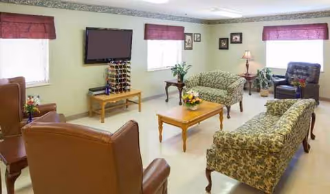 Bright common living room with multiple couches, tables, plants, and a wall-mounted TV.