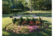 A well-maintained outdoor garden area with a circular flower bed filled with pink and red flowers, surrounded by neatly trimmed bushes. In the background, there are trees, a grassy lawn, and benches along a paved pathway.
