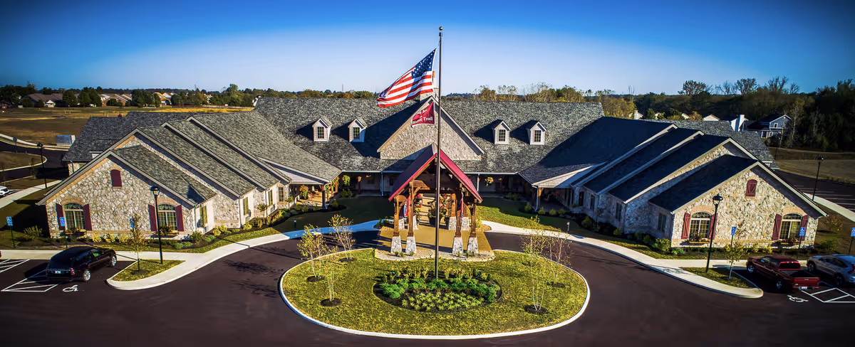 Aerial view of The Inn at Walnut Trail, a large single-story stone building with a gray shingled roof and multiple dormer windows. In front of the building is a circular driveway with a landscaped island featuring a flagpole flying the American flag and a facility flag. Several cars are parked in designated parking spaces around the building. The surrounding area includes open fields and trees under a clear blue sky.