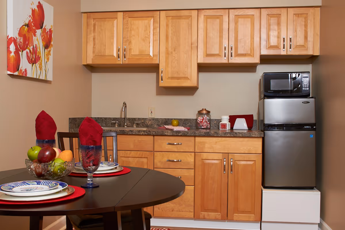A small kitchen area with wooden cabinets, a countertop with a sink, a microwave on top of a mini refrigerator, and a round dining table set with plates, glasses with red napkins, and a bowl of fruit. A floral painting is hung on the wall to the left.