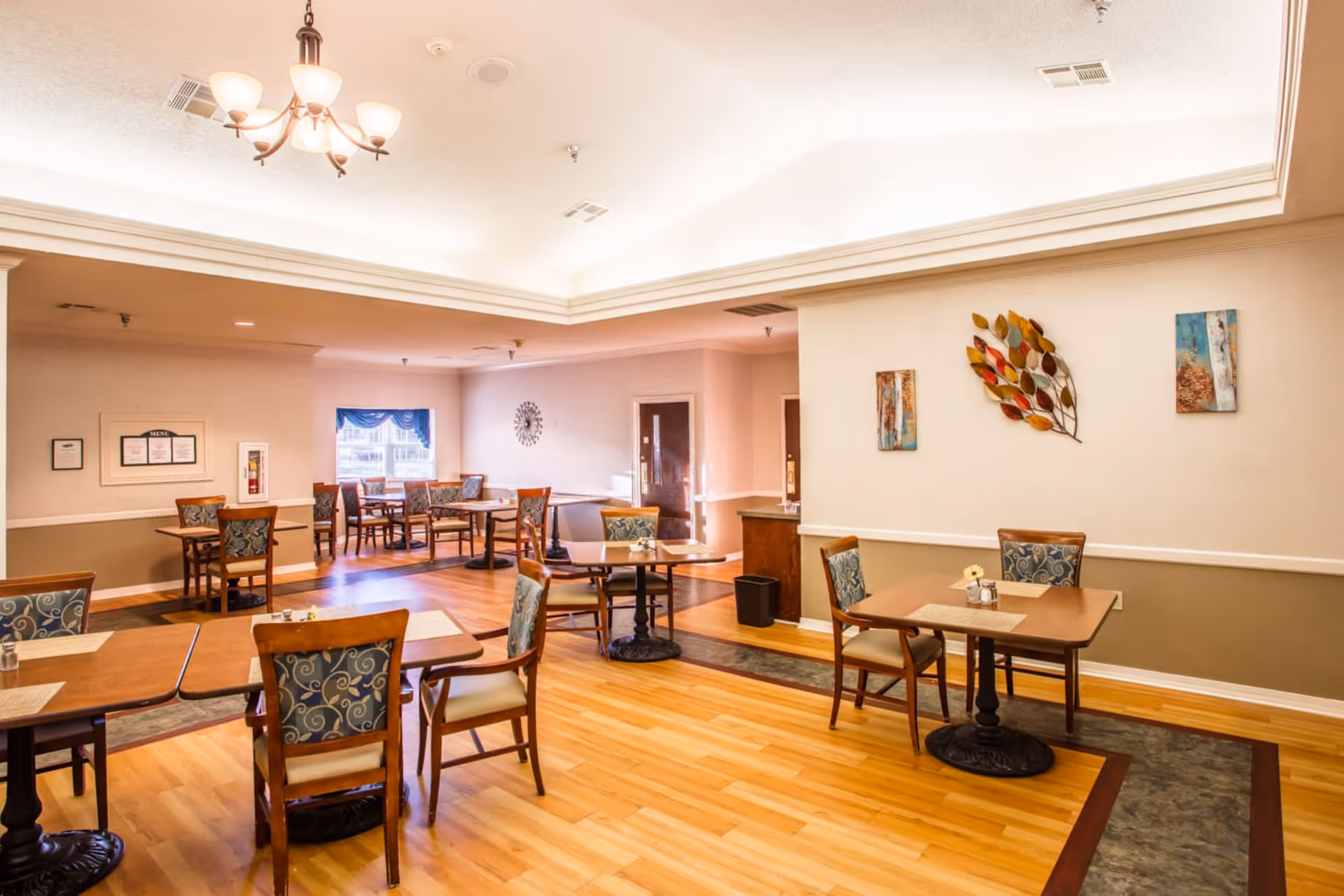 A well-lit dining room with wooden floors and multiple tables and chairs arranged neatly. The walls are decorated with colorful artwork, and a chandelier hangs from the ceiling. There is a window with blue curtains and a clock on the far wall.