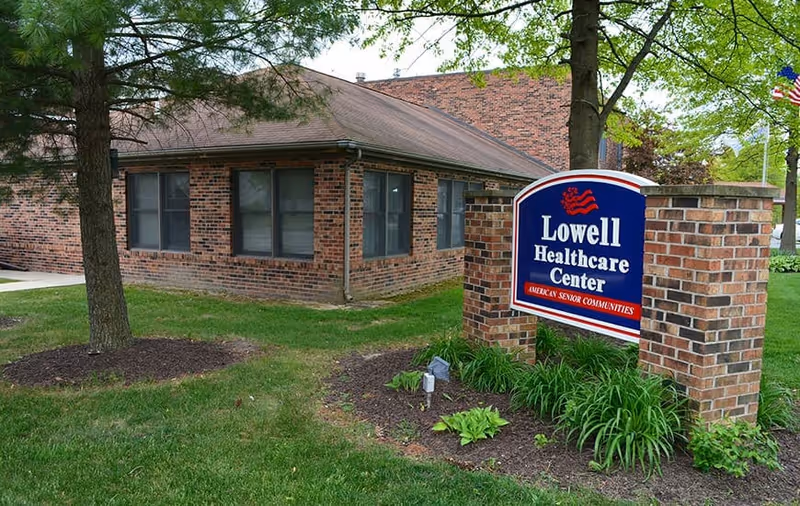 Exterior view of a brick building with multiple windows, surrounded by green grass and trees. In front of the building is a blue and red sign that reads 'Lowell Healthcare Center American Senior Communities.'