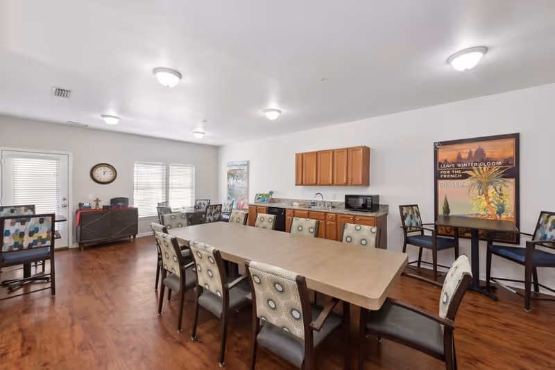 A spacious dining room with a long rectangular table surrounded by chairs with patterned upholstery. The room has wooden flooring, white walls, and ceiling lights. There is a kitchenette area with wooden cabinets, a microwave, and a sink. Two large posters are on the walls, and there are additional tables and chairs near the windows and door.