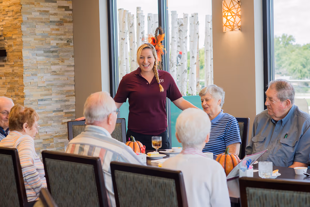A group of elderly people sitting around a dining table with a smiling staff member standing and engaging with them in a well-lit room with large windows and decorative birch branches outside.