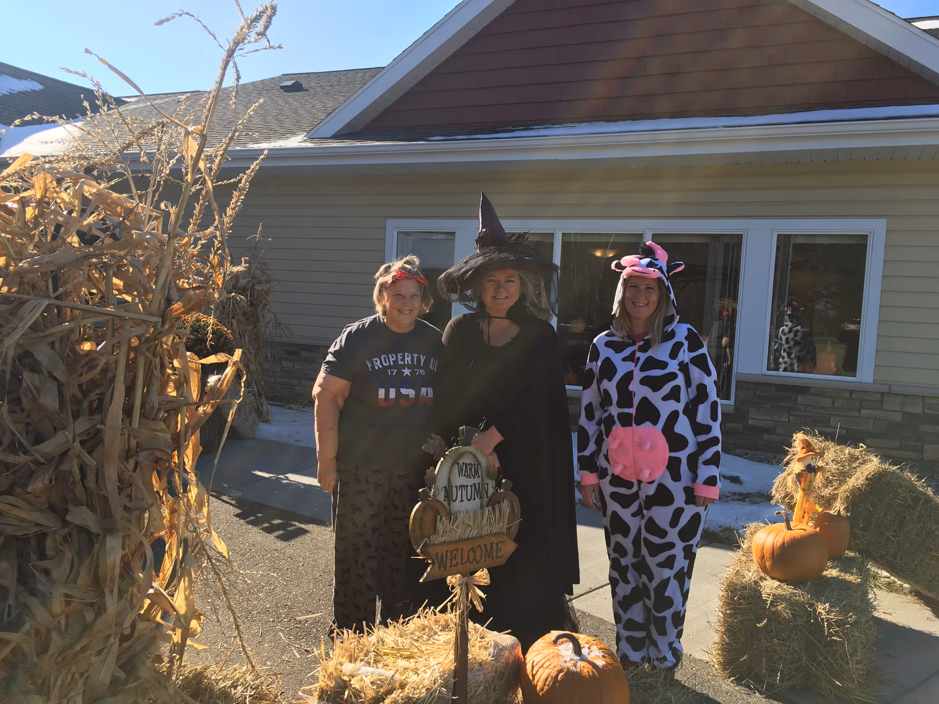Three women standing outside a building decorated for autumn. One woman is wearing a black witch costume, another is dressed in a cow costume, and the third is wearing casual clothes with a red headband. There are pumpkins, hay bales, and dried cornstalks around them, with a sign that says 'Warm Autumn Welcome'.