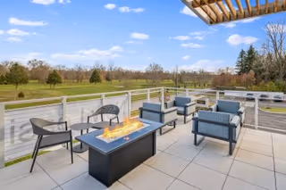 Rooftop outdoor seating area with modern chairs, a rectangular fire pit table, and a view of grassy grounds and trees.