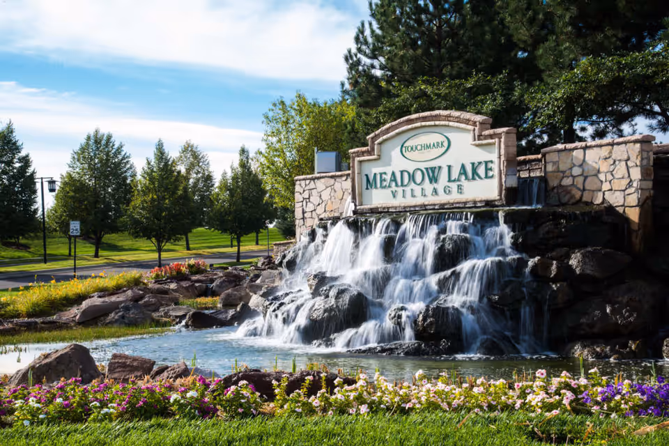 A landscaped outdoor area featuring a stone waterfall with water cascading over rocks into a pond, surrounded by colorful flowers and greenery. Behind the waterfall is a stone sign that reads 'Touchmark Meadow Lake Village'. Trees and a road with a speed limit sign are visible in the background under a partly cloudy sky.