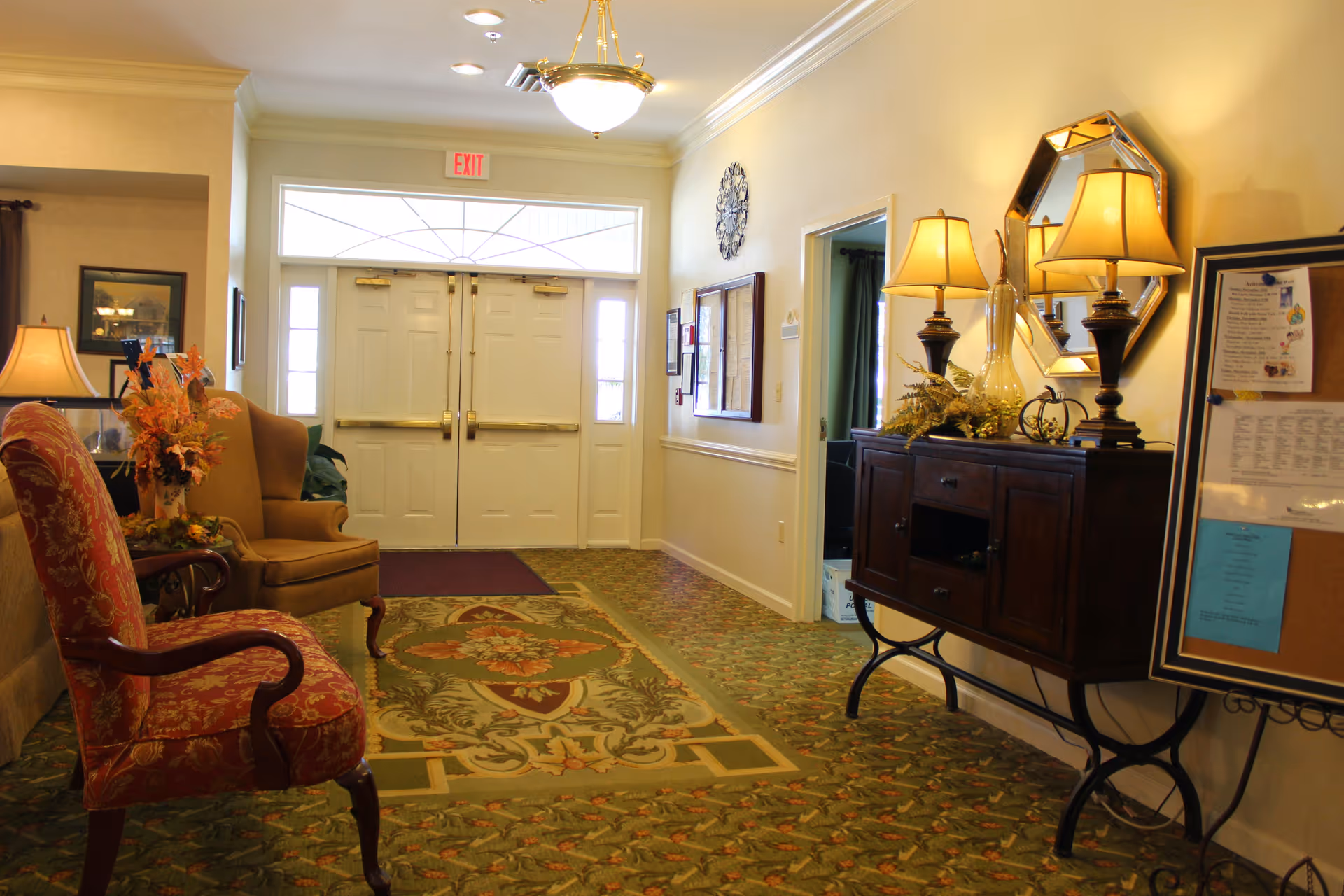 Well-lit senior living entrance lobby with upholstered chairs, a console table with lamps, decorative rug, and double exit doors.