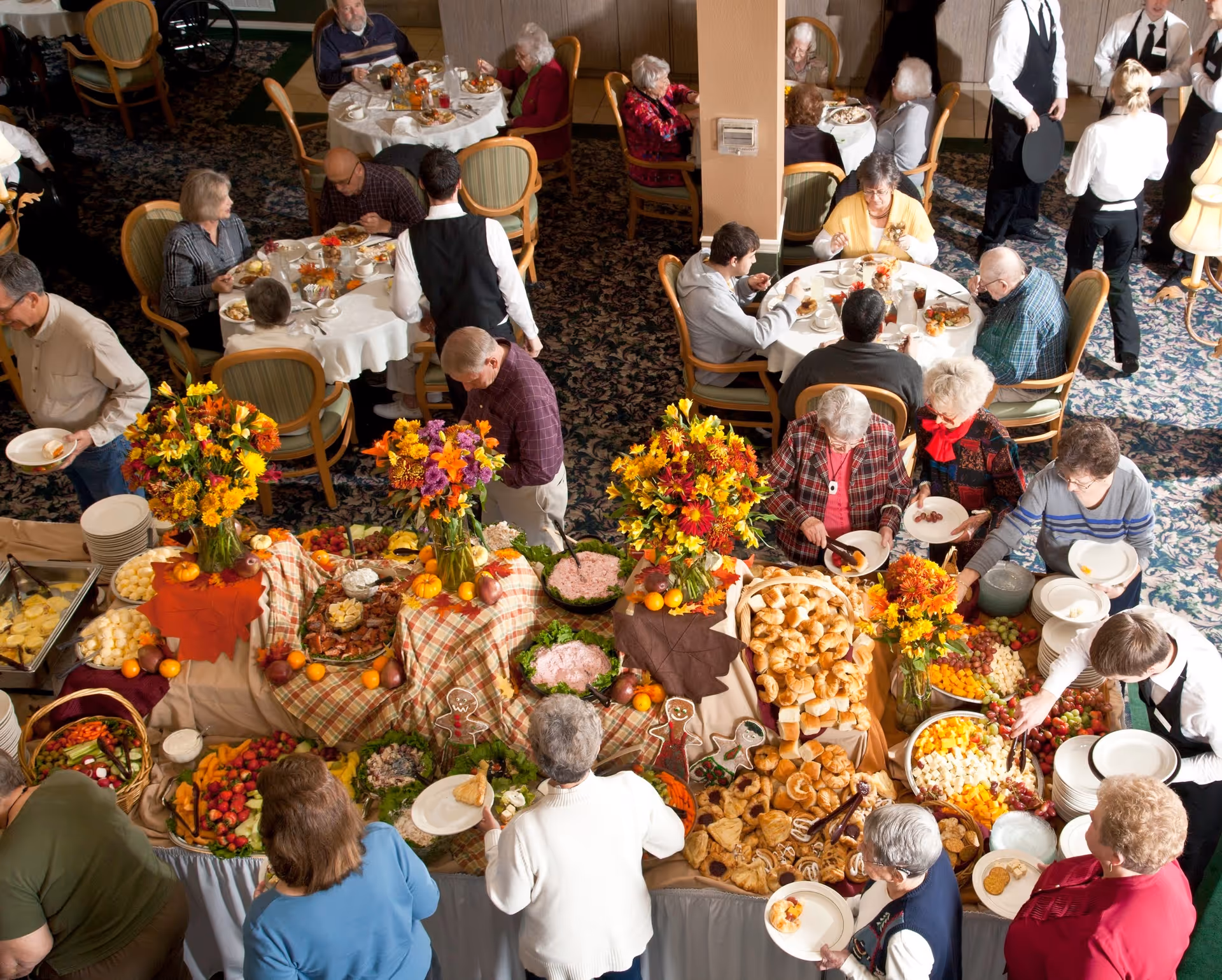Overhead view of seniors and servers gathered around a large buffet table filled with pastries, fruit, and floral centerpieces in a dining room.