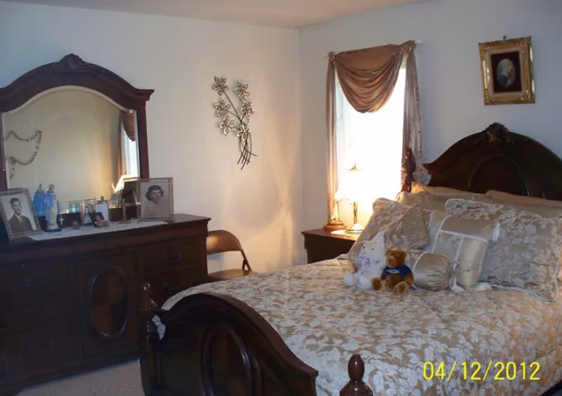 A bedroom with a dark wooden bed frame and matching dresser with a large mirror. The bed is made with a beige floral comforter and several decorative pillows, along with two stuffed animals placed on top. A nightstand with a lamp is next to the bed, and a window with draped curtains lets in natural light. There are framed photos on the dresser and a decorative wall piece on the wall.