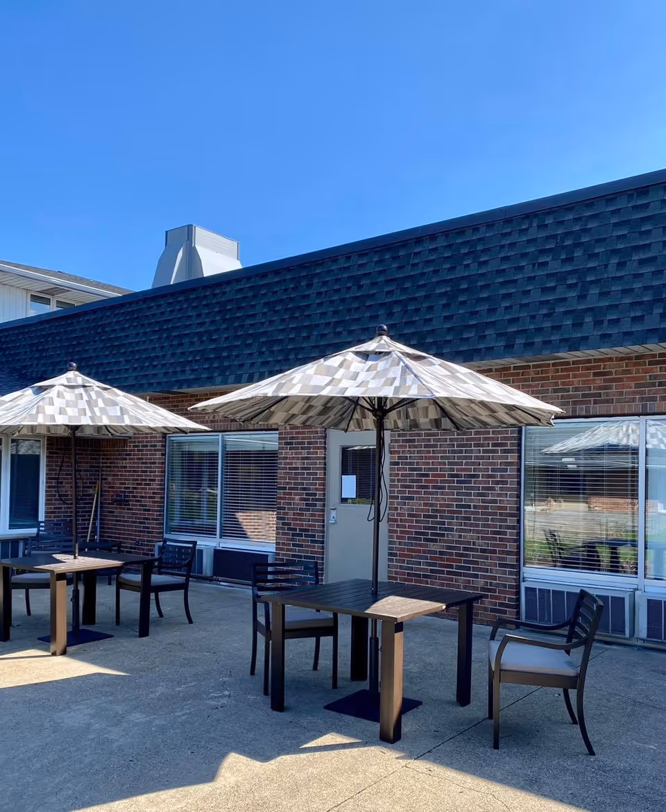 Outdoor patio area with two tables, each shaded by a large umbrella. Each table is surrounded by chairs. The patio is adjacent to a brick building with windows and a door. The sky is clear and blue.