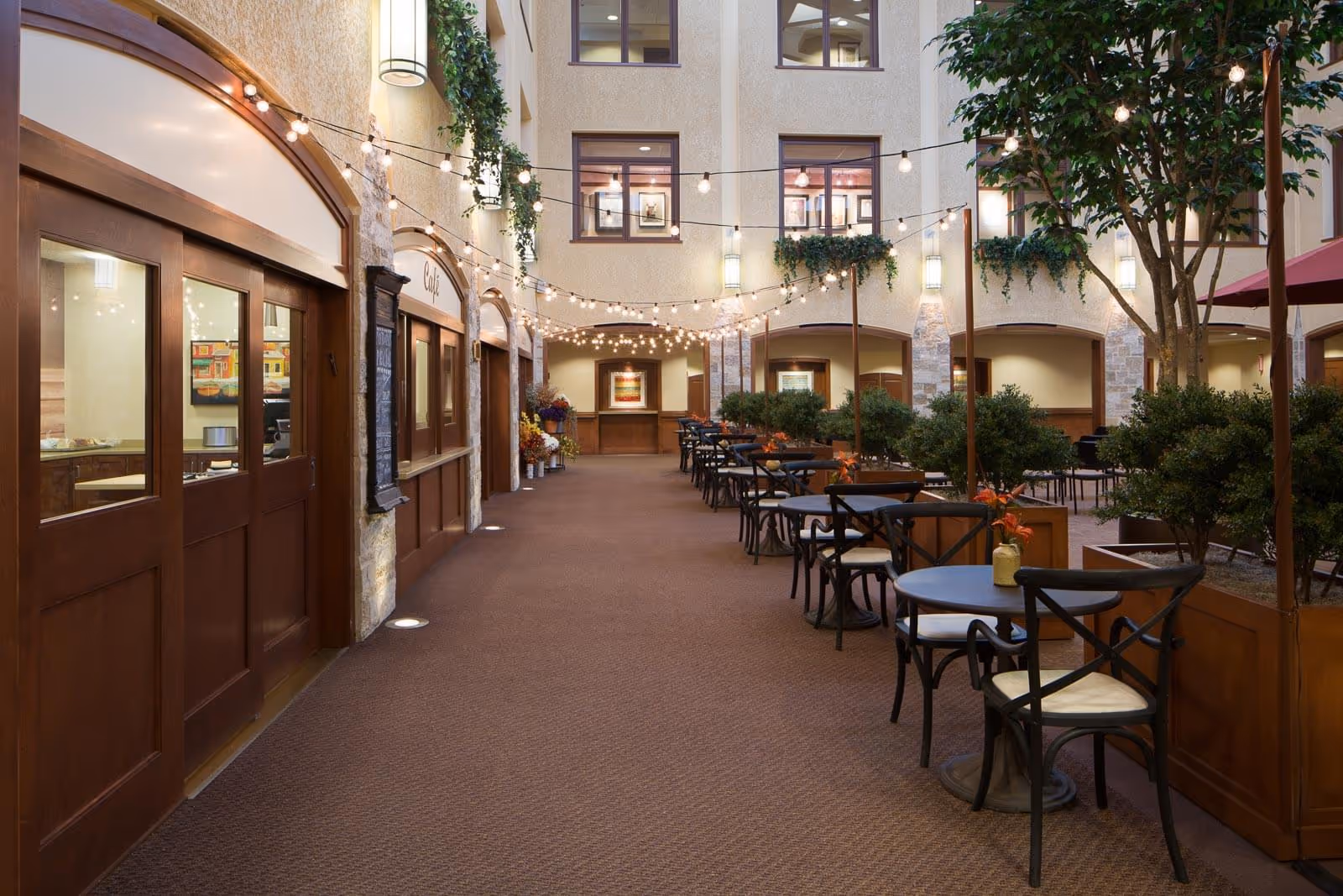 Indoor atrium-style dining area with tables and chairs, string lights, potted trees, and a café counter.